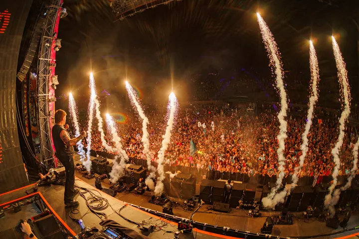 Seen from the back of the stage, a performer at the edge of the deck facing a full crowd as pyro jets arc over the barricade.