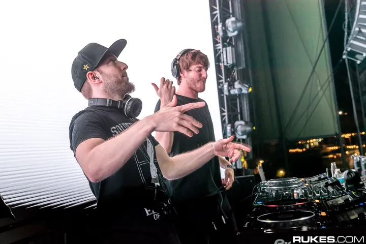 Two DJs behind the decks at dusk, one gesturing to the crowd, stage scaffolding and the darkening sky behind them.