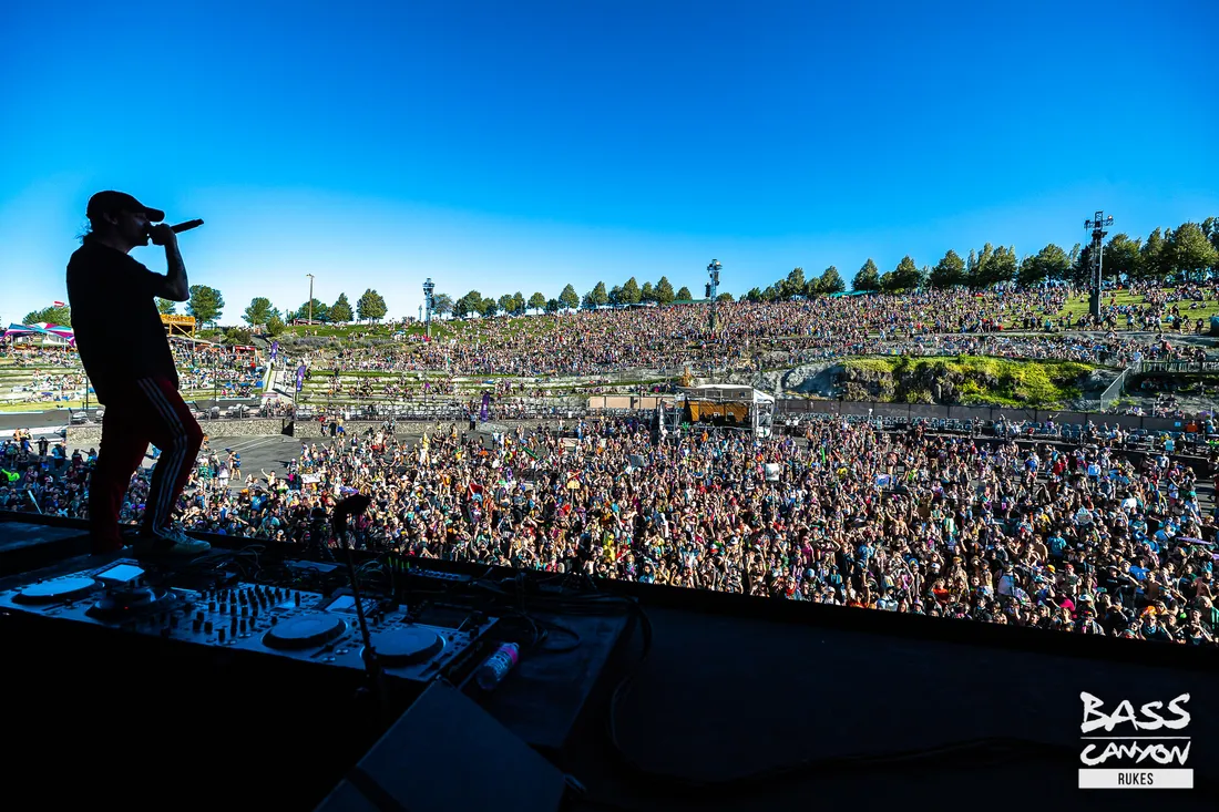 Seen from behind the decks, a performer facing the Gorge's full grass slope packed with people in daylight, the far cliffs and treeline beyond.
