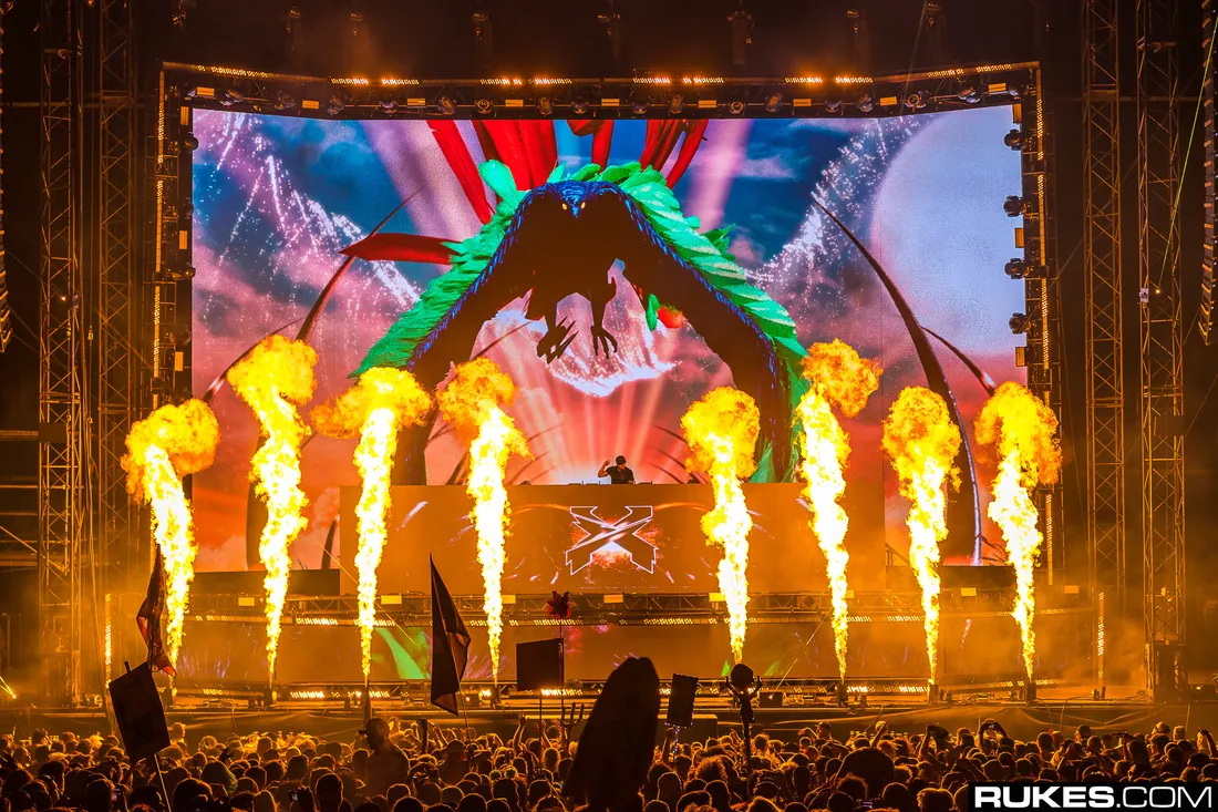 The Paradox stage at night, eight flame jets firing across the front of the deck under a wide LED wall, the crowd silhouetted below.