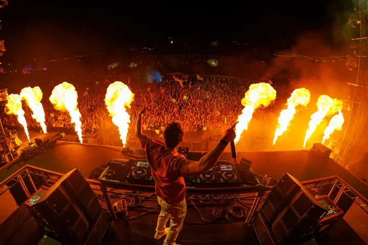 A DJ seen from behind at the decks with both arms up, six flame jets firing across the front of the stage at night.